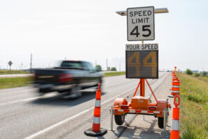 Black truck driving on a highway past a speed limit sign next to orange caution road cones.